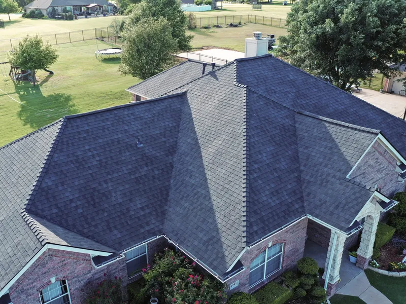 Aerial view of a single-story brick ranch home with a finished charcoal shingle roof and new gutters, a stone entry column at the front and a back yard with mature trees beyond.
