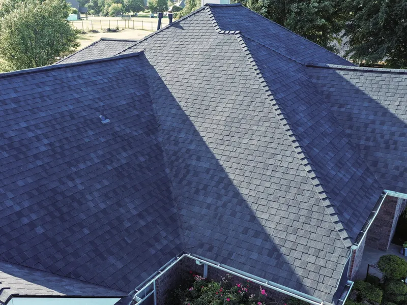 Close aerial view of a finished asphalt shingle roof, showing several hip and ridge lines with ridge cap installed, clean shingle courses, white gutters along the eaves and flowering shrubs below.