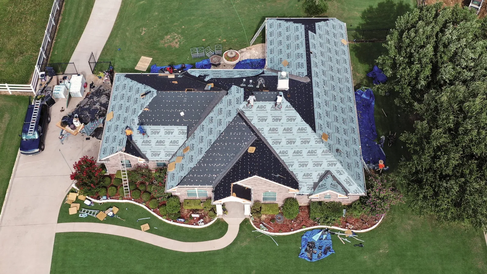 Overhead view of a large brick home mid-replacement, with most roof slopes stripped to the decking and covered in synthetic underlayment, shingle bundles staged on the ridges and tarps protecting the landscaping below.