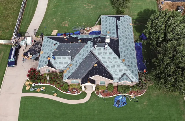 Overhead view of a large brick home mid-replacement, with most roof slopes stripped to the decking and covered in synthetic underlayment, shingle bundles staged on the ridges and tarps protecting the landscaping below.