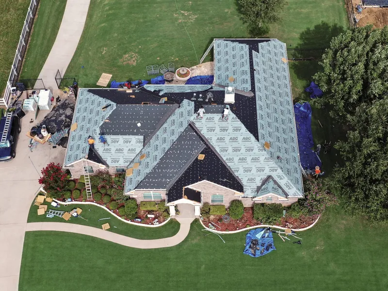 High overhead view of a large brick home with every roof slope stripped and covered in felt and synthetic underlayment, blue tarps laid over the patio furniture and landscaping below, a stone fountain and a circular drive.