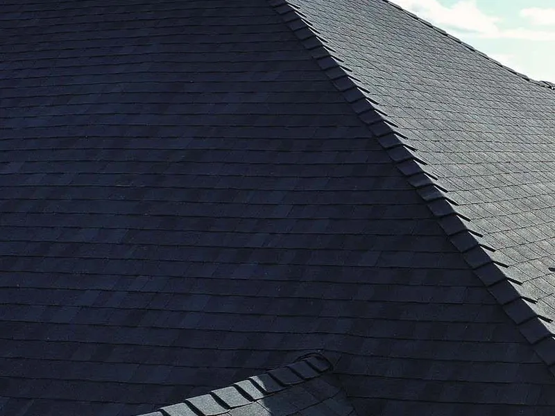Upward view from the ground of a finished dark architectural shingle roof against a blue sky, showing the hip line, the ridge cap, a sided chimney where it meets the shingles, and a brick gable with an exterior light fixture.