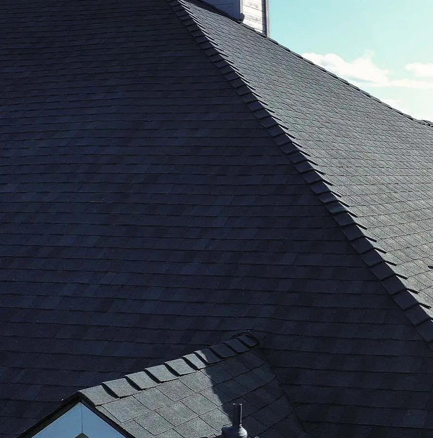 Close view of a finished dark architectural shingle roof against a blue sky, showing the hip line, ridge cap and a sided chimney where new flashing meets the shingles.