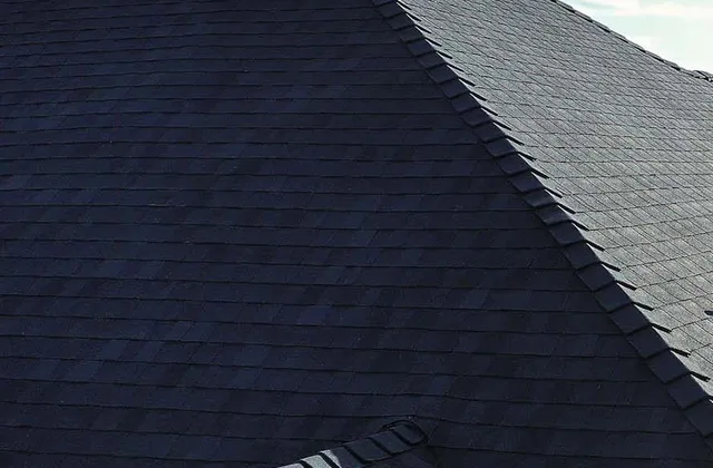 Close view of a finished dark architectural shingle roof against a blue sky, showing the hip line, ridge cap and a sided chimney where new flashing meets the shingles.