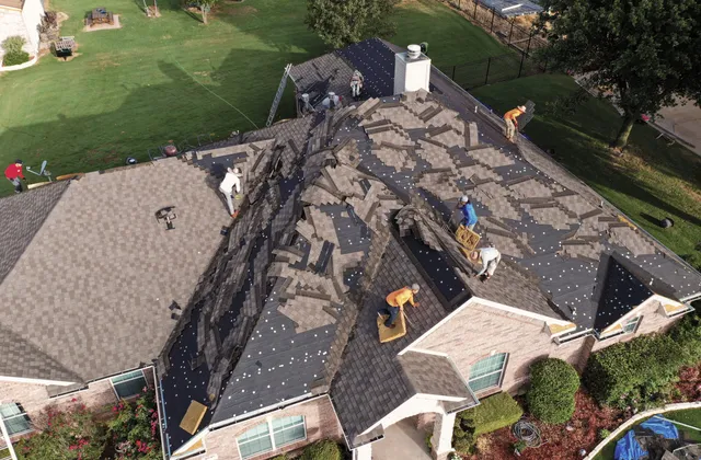 Aerial view of a roofing crew stripping storm-damaged shingles off a large hip roof, with rows of removed shingles piled across the slopes, exposed decking and felt, and ladders set at the eaves.