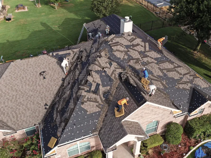 Aerial view of a roofing crew stripping the old shingles from a large hip roof, with removed shingles piled in rows across the slopes, sections of bare decking and felt exposed, and ladders set at the eaves.