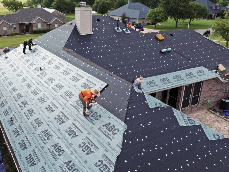 Two roofers rolling out synthetic underlayment across a stripped roof slope, one in an orange shirt and straw hat and one in a green shirt, with black felt on the adjacent slope and a brick ranch house in the background.