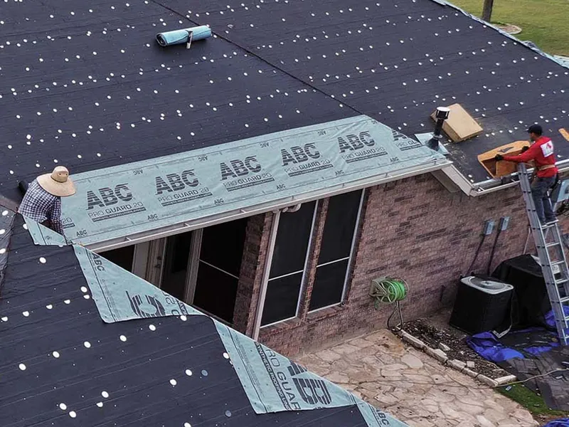 Aerial view of a home built around an open courtyard, its roof stripped and covered in black felt and synthetic underlayment, with crew members working near the ridge, a ladder at the eave and a stone patio below.